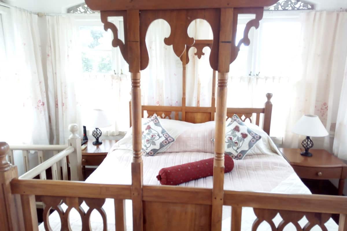 A four-poster bed with a wooden frame, a bedside table with a lamp, and a window with floral curtains.