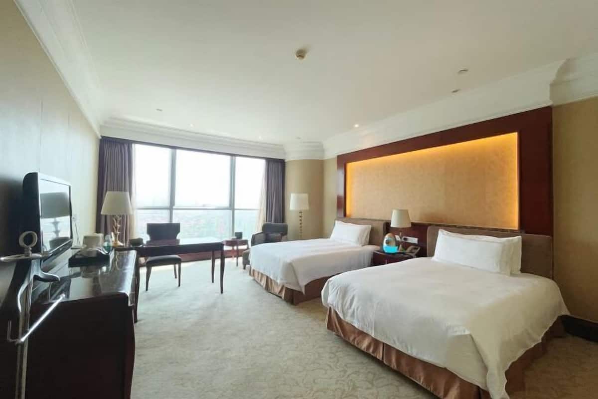 A hotel room with two beds, a desk with a TV, and a large window offering a view.