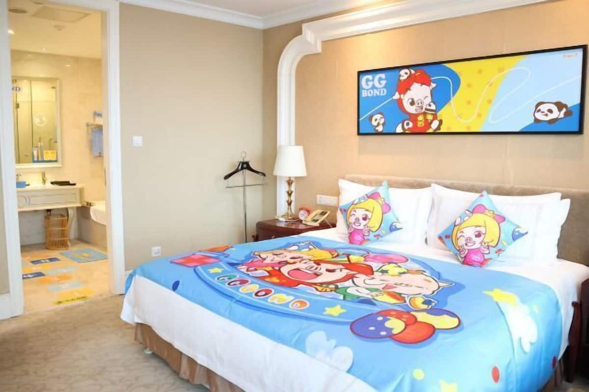 A hotel room with a bed featuring cartoon character bedding, a television mounted on the wall, and a bathroom visible through an open door.