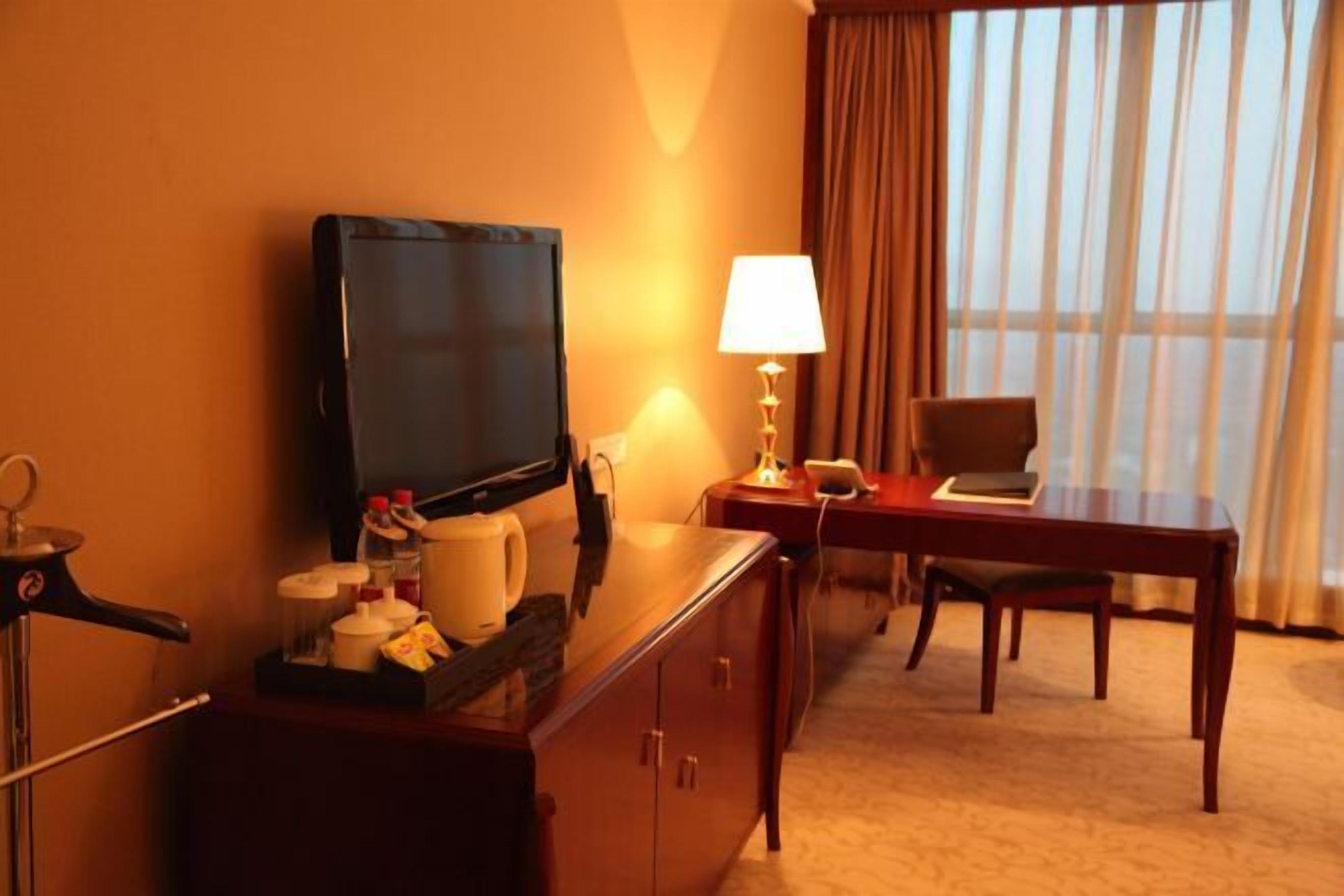 A hotel room with a TV, a desk with a lamp, a chair, and a window with curtains.