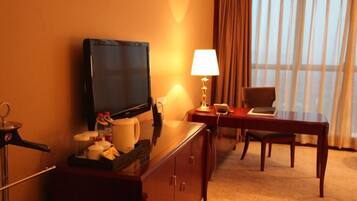 A hotel room with a TV, a desk with a lamp, a chair, and a window with curtains.