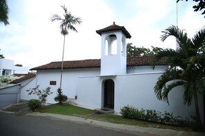 A white building with a bell tower, a tiled roof, and a small garden with palm trees.