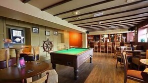 Une table de billard se trouve dans une pièce dotée d’un plancher en bois et d’un plafond haut avec des poutres apparentes. On y voit plusieurs chaises et tables en bois, ainsi qu’un coin bar à l’arrière-plan.