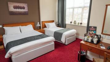 A hotel room with two single beds, a wooden headboard, a desk with a tray of breakfast items, a trash can, and a window with curtains.
