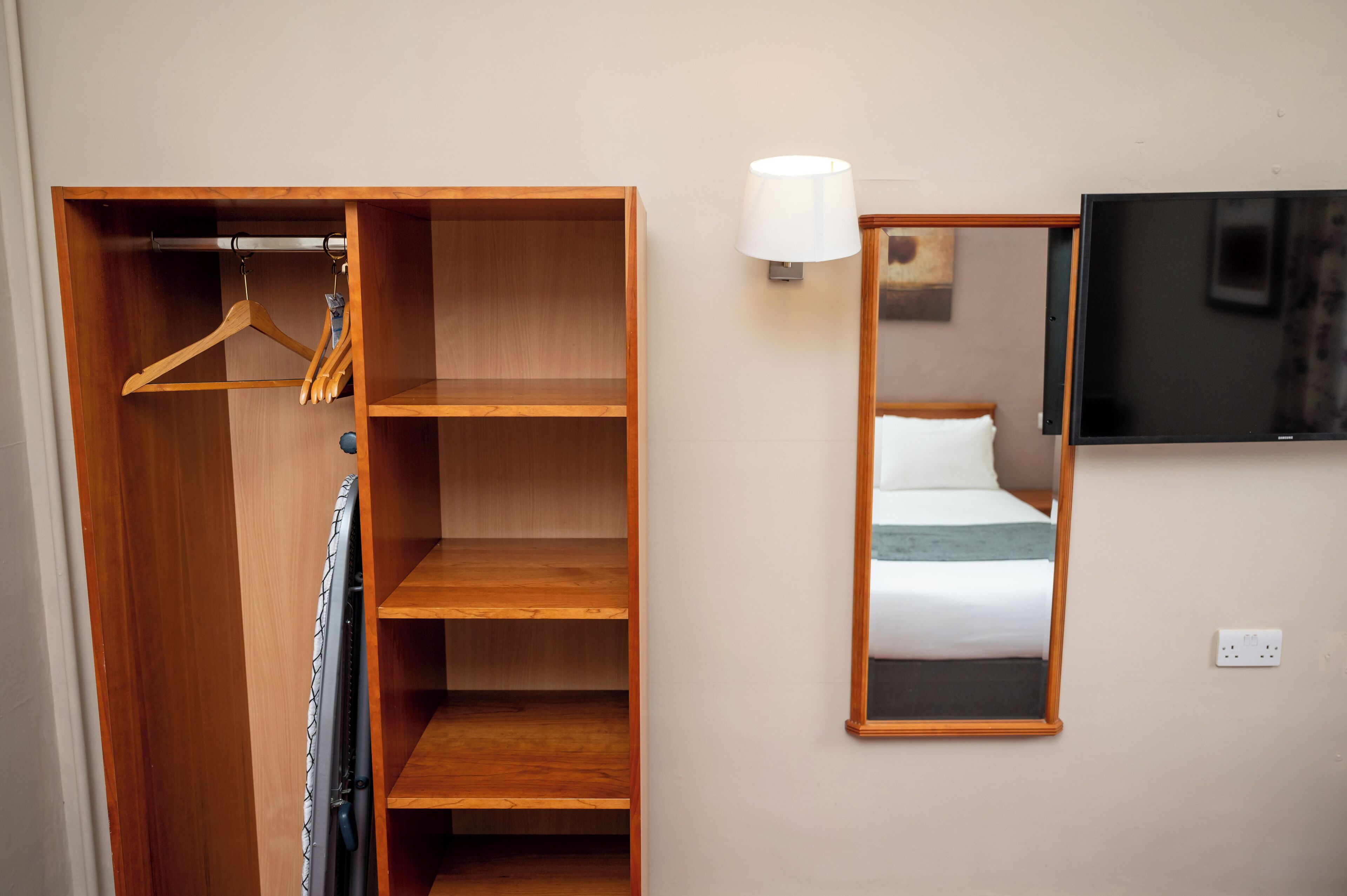 Une chambre d’hôtel avec une armoire en bois, un miroir, un lit recouvert d’une couverture blanche et turquoise, et un téléviseur à écran plat fixé au mur.