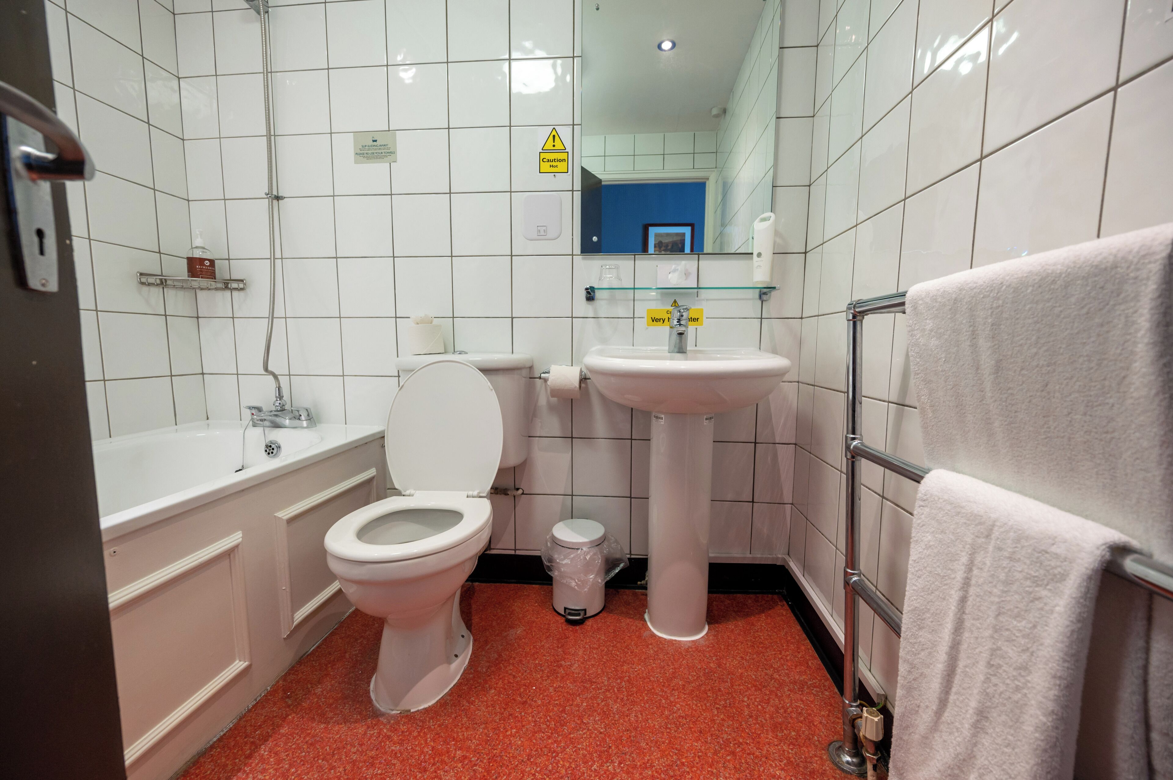 Un baño con inodoro, lavabo y bañera. Hay un letrero de precaución en la pared, un cubo de basura y un perchero para toallas.
