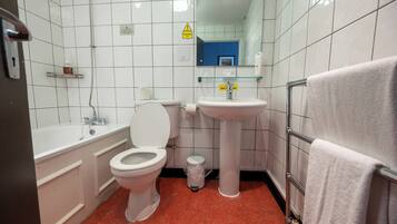 Un baño con inodoro, lavabo y bañera. Hay un letrero de precaución en la pared, un cubo de basura y un perchero para toallas.
