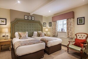 A bedroom with two beds, a nightstand, a chair, and a window with curtains. - The Royal Oak (Welshpool)