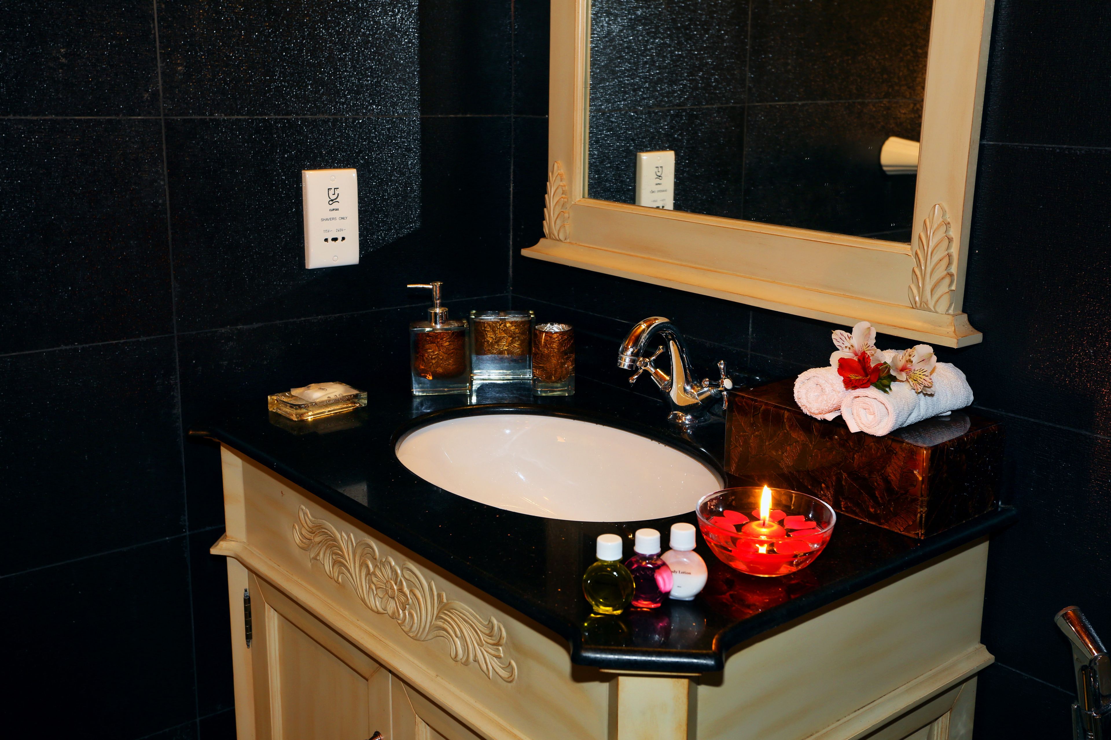 A bathroom with a black countertop, white sink, and a mirror above. There are toiletries on the counter, including a candle, bottles, and a soap dish.