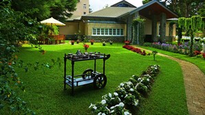 A well-maintained garden with a cart of tea set, a lawn, and a building with a stone facade. - The Firs (Nuwara Eliya)