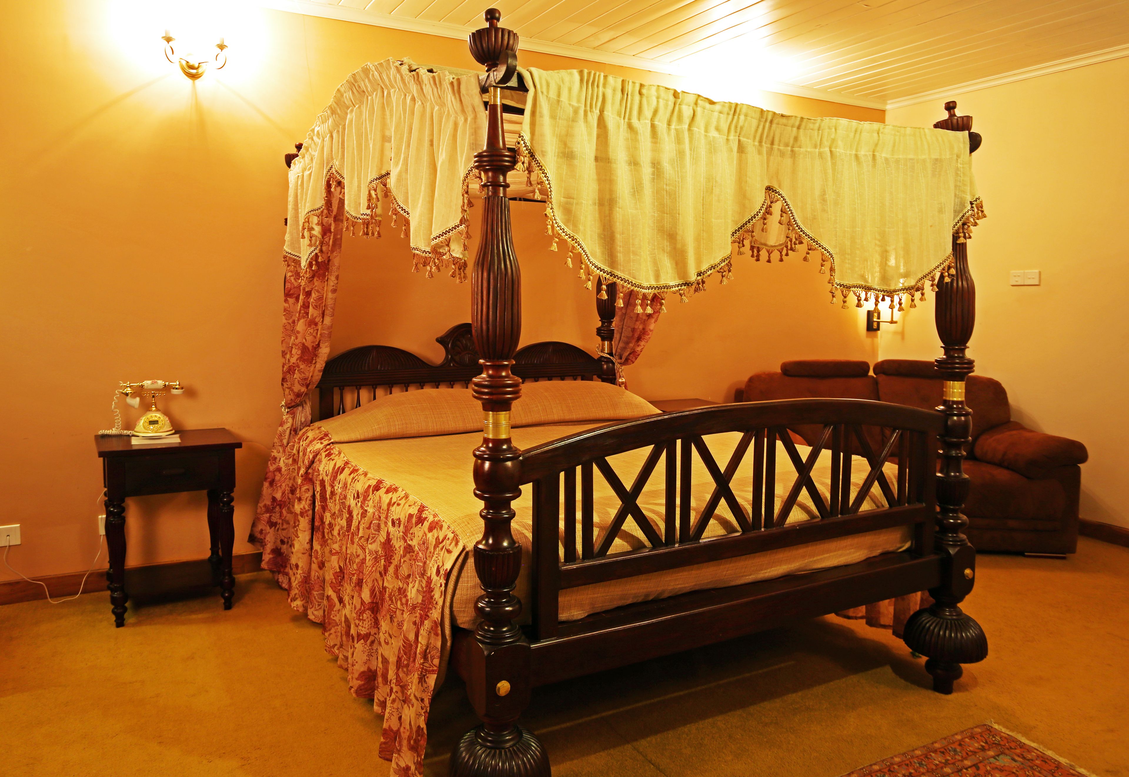 Uma cama de dossel com cortina florida, um criado-mudo de madeira com abajur e uma cadeira em um quarto com iluminação acolhedora.