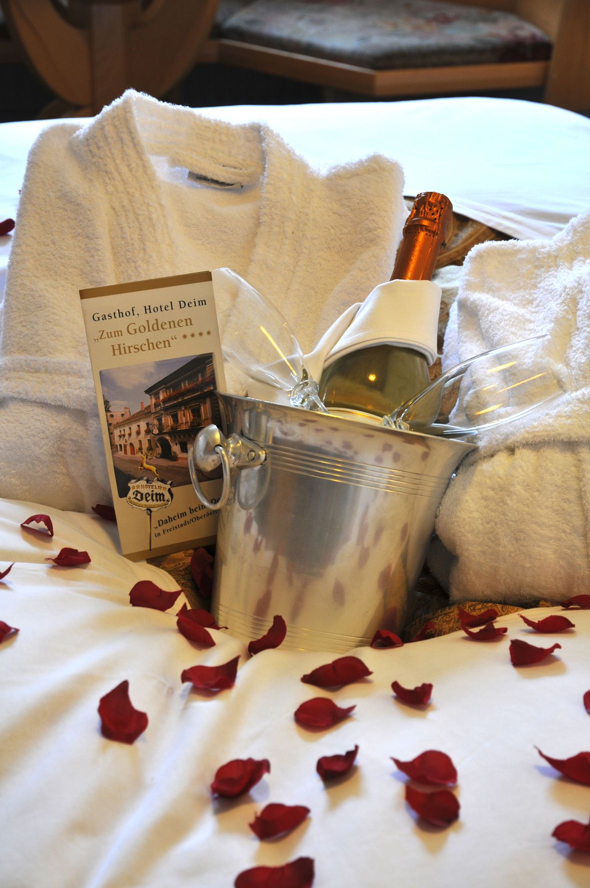A bed with rose petals, a bottle of champagne, and a book titled "Zum Goldenen Hirschchen".