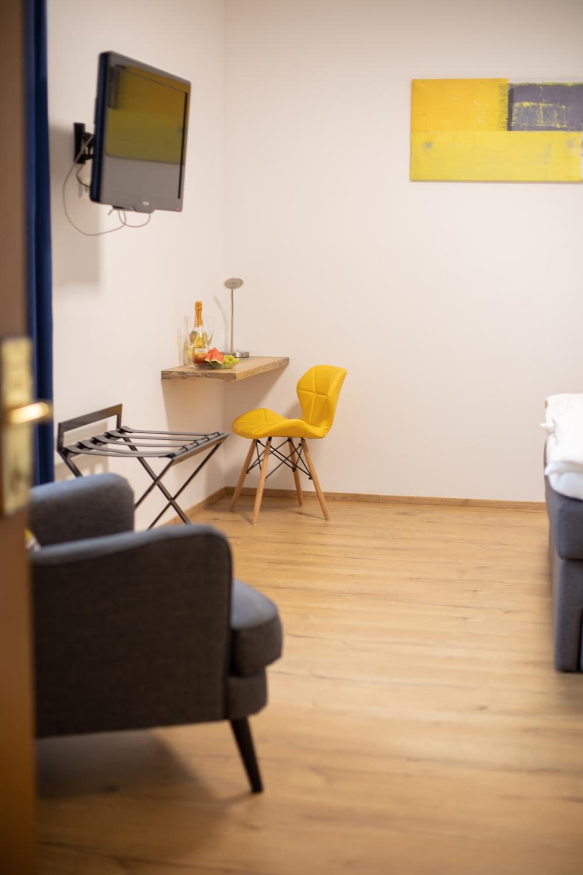 Una habitación con piso de madera, un sofá gris, un sillón amarillo, una mesita con una botella y un televisor montado en la pared.