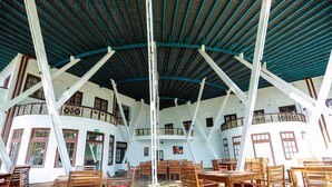 A spacious indoor area with multiple wooden tables and chairs, a high ceiling with a blue and white color scheme, and a view of the outside through large windows. - Welcombe Hotel (Trincomalee)