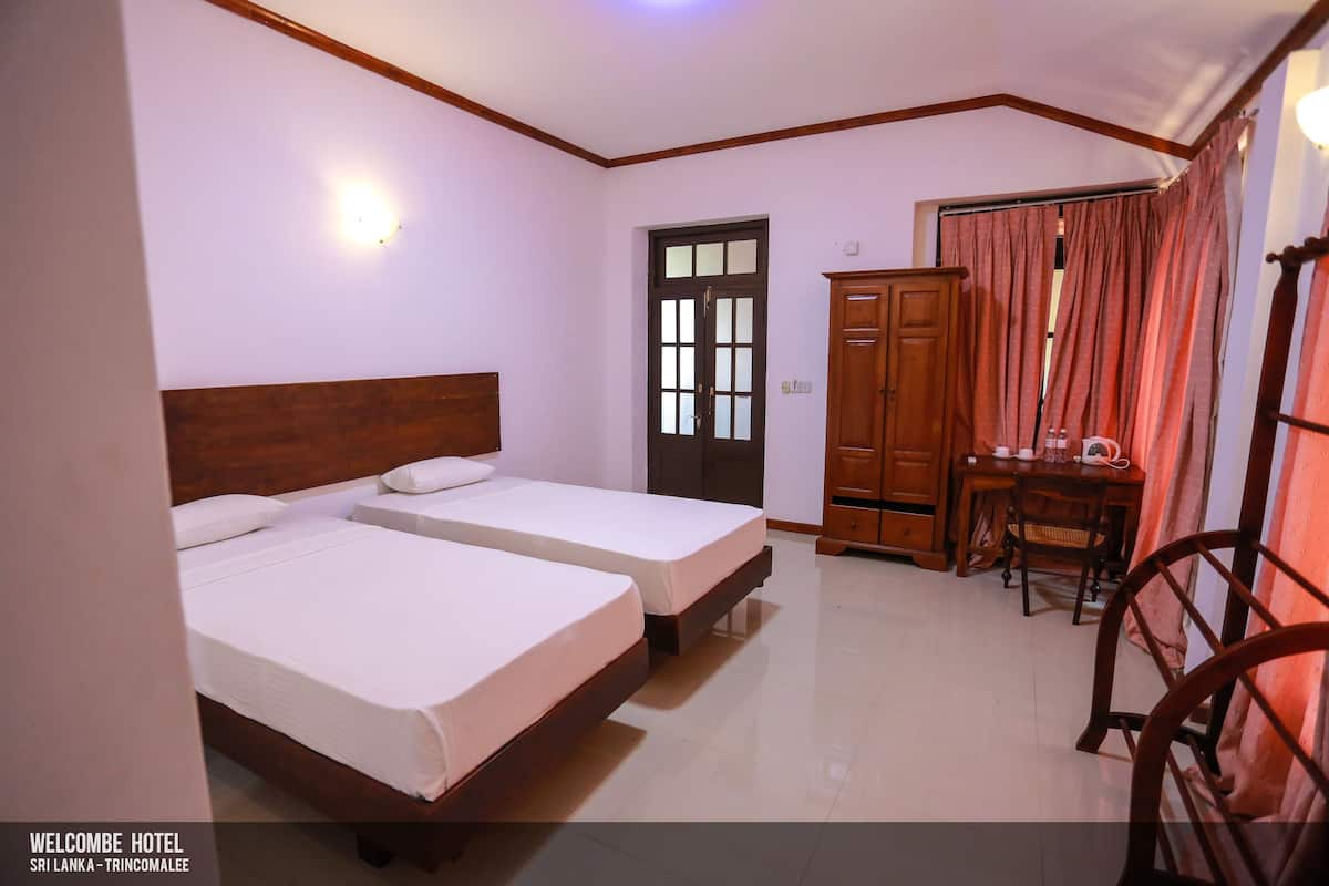 A hotel room with two single beds, a wooden wardrobe, a desk with a chair, and a window with curtains.