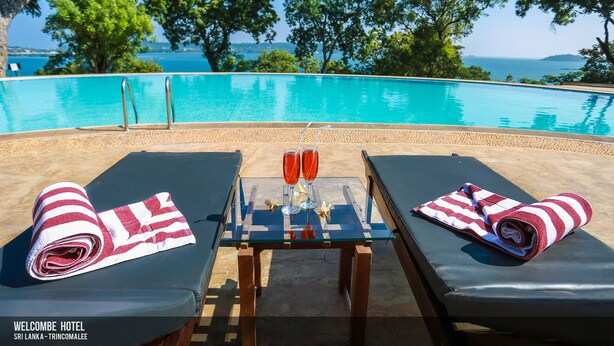 A poolside setting with lounge chairs, a table with towels and drinks, and a view of the sea. - Welcombe Hotel (Trincomalee)