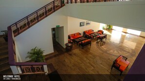 A hotel lobby with a wooden staircase, seating area with orange sofas and tables, and a large window providing natural light. - Welcombe Hotel (Trincomalee)