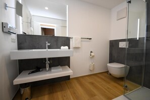 Ein modernes Badezimmer mit einem weißen Waschbecken, dunkelgrauen Wandfliesen, einer Duschkabine aus Glas und Holzboden.