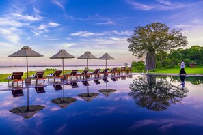 A poolside area with lounge chairs and umbrellas, a tree, and a clear sky. - EKHO Lake Polonnaruwa (Polonnaruwa)