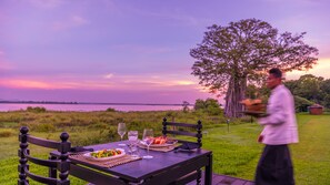 A dining table set outdoors with a view of a lake and trees at sunset. - EKHO Lake Polonnaruwa (Polonnaruwa)