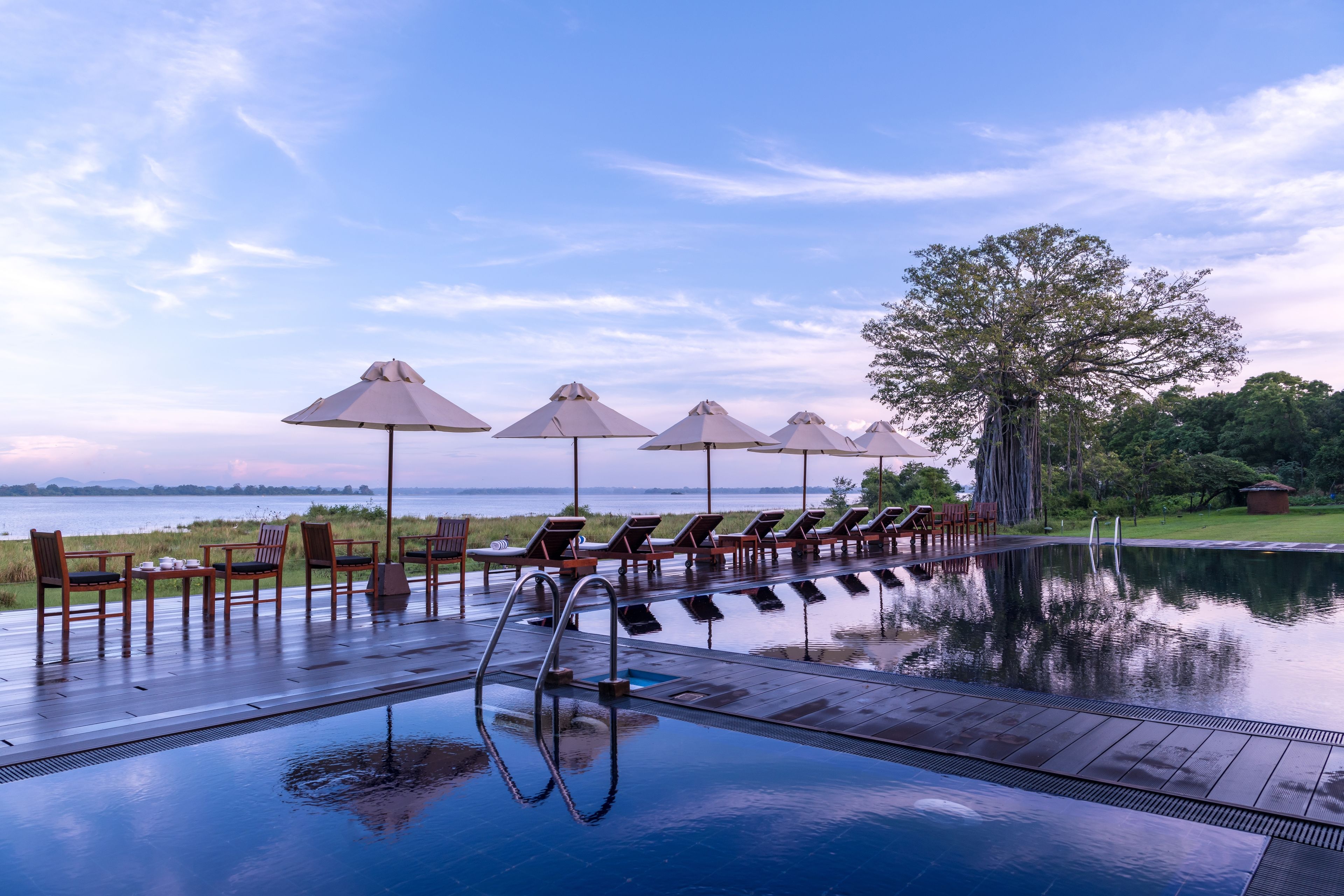 An infinity pool with lounge chairs and umbrellas, surrounded by a wooden deck and a view of the water.