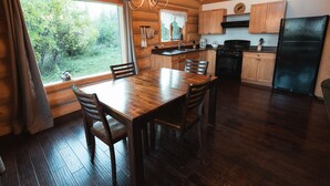 A wooden cabin interior with a dining table, chairs, a chandelier, a kitchen with wooden cabinets, and a large window overlooking greenery. - Ten-ee-ah Lodge & Campground (Lac La Hache)