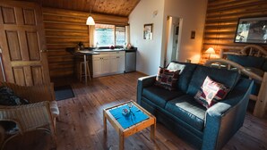 A cozy cabin interior with a wooden ceiling, a dining area with a table and chairs, a sofa with cushions, and a kitchen with a window. - Ten-ee-ah Lodge & Campground (Lac La Hache)