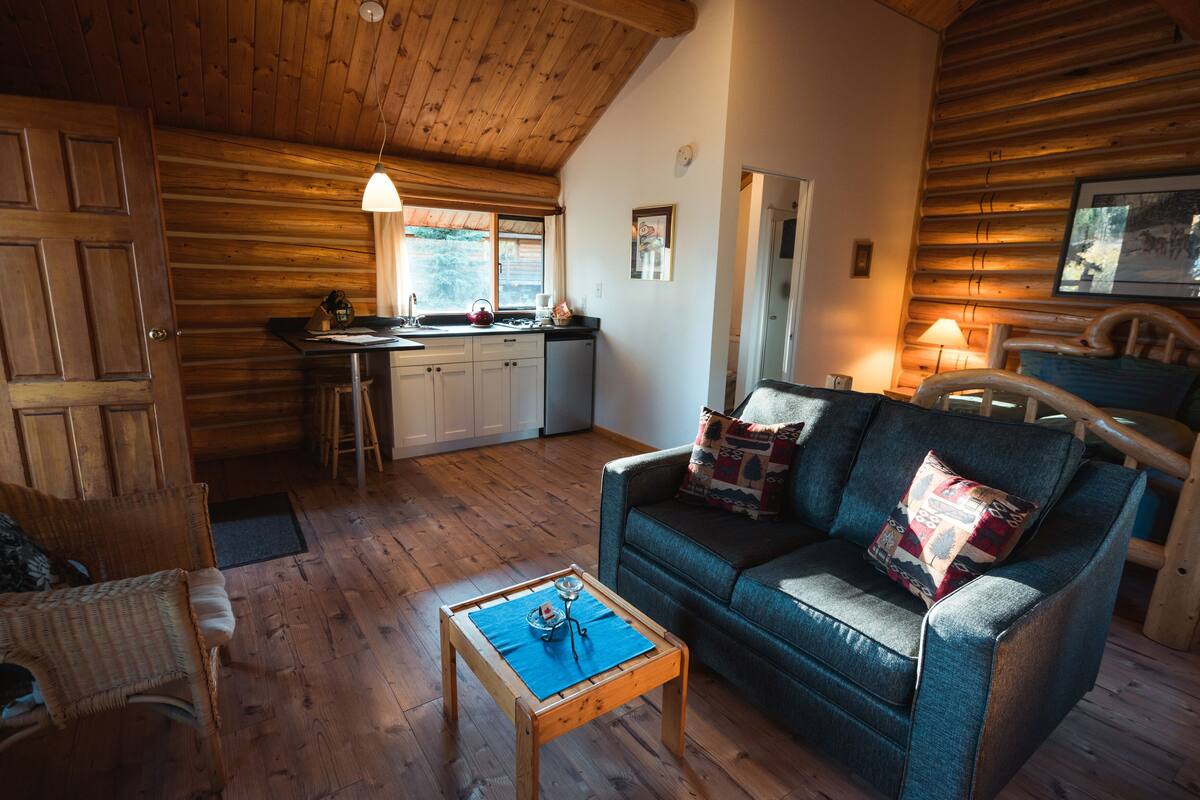Un interior de cabaña acogedor con techo de madera, una zona de comedor con mesa y sillas, un sofá con cojines y una cocina con ventanal.