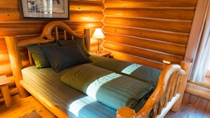 A wooden cabin bedroom with a bed, pillows, a nightstand, a lamp, and a framed picture on the wall. - Ten-ee-ah Lodge & Campground (Lac La Hache)