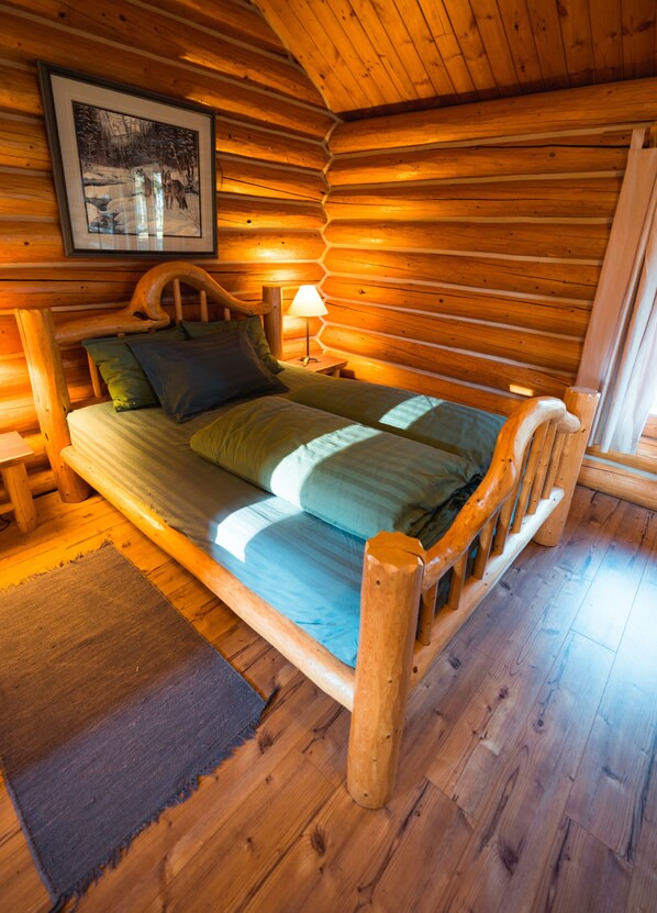 A wooden cabin bedroom with a bed, pillows, a nightstand, a lamp, and a framed picture on the wall. - Ten-ee-ah Lodge & Campground (Lac La Hache)