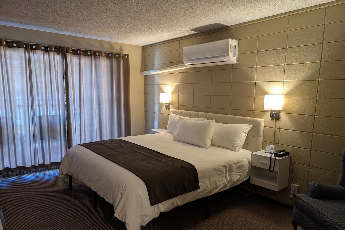 A hotel room with a bed, bedside lamps, an air conditioning unit, a nightstand, and a sofa.