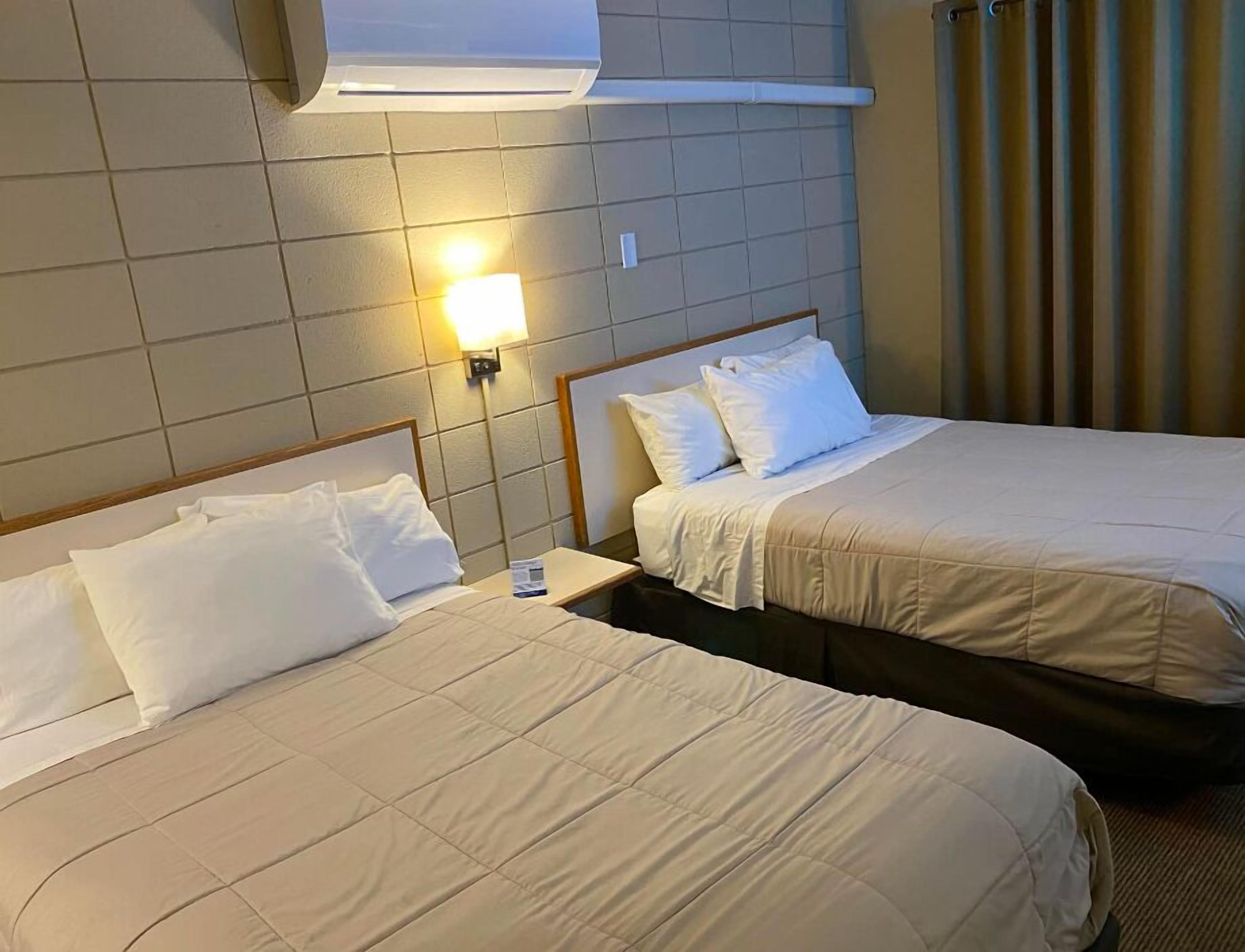 A hotel room with two beds, each with white linens and pillows. There is a nightstand with a lamp between the beds.