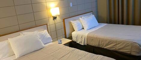 A hotel room with two beds, each with white linens and pillows. There is a nightstand with a lamp between the beds.