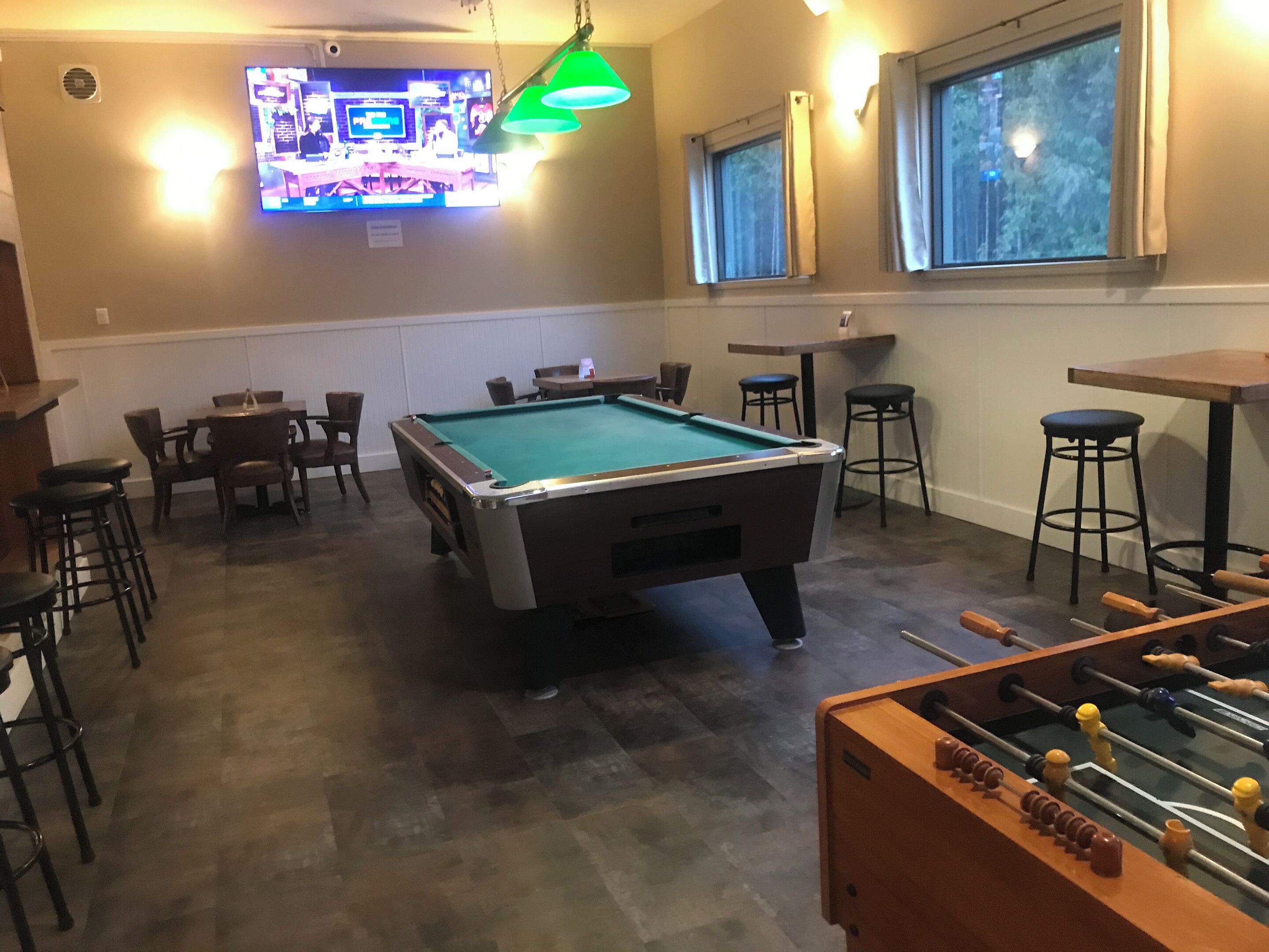 Une table de billard dans une pièce où est fixée une télévision au mur, ainsi qu’une table de baby-foot, plusieurs chaises et des tabourets.