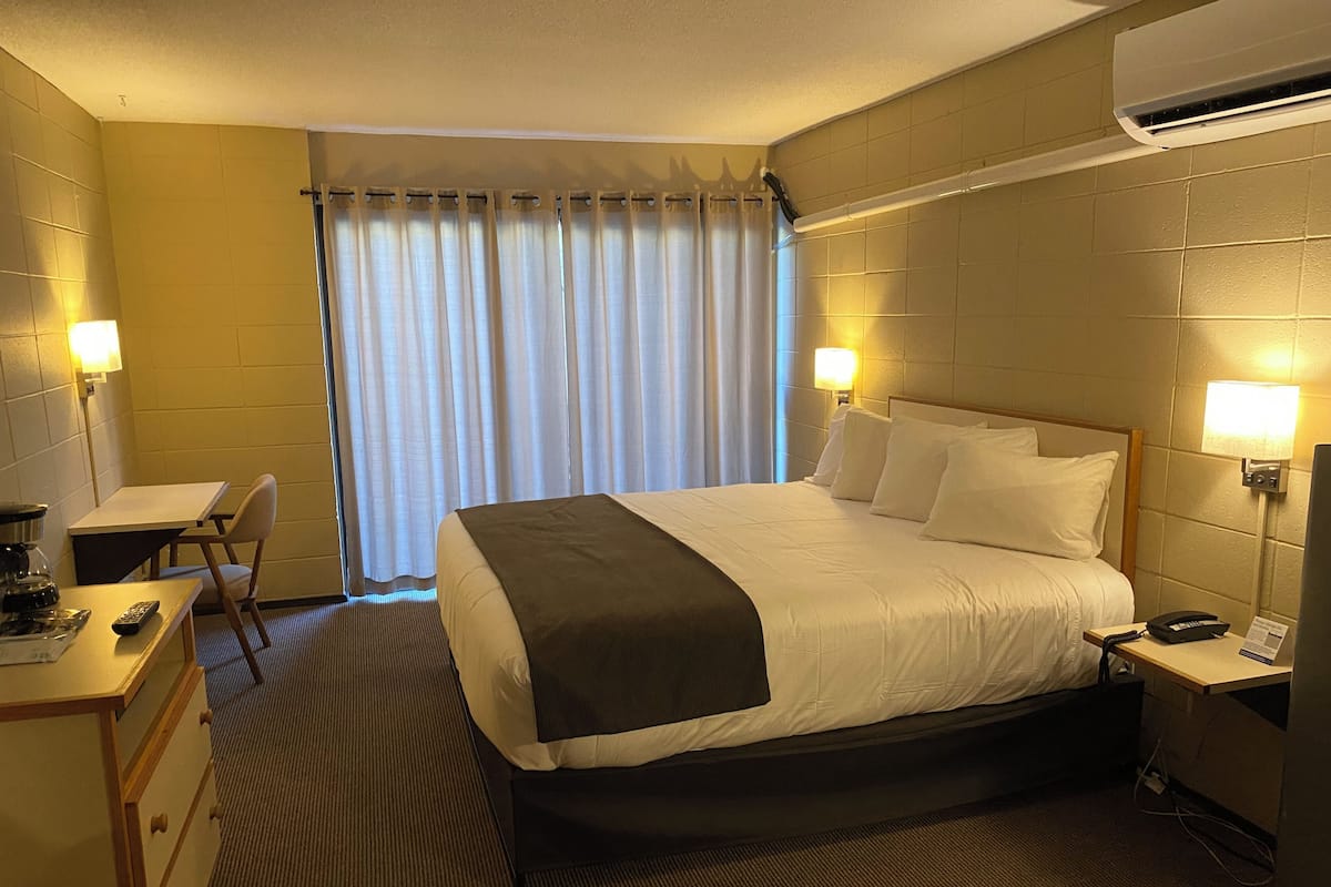 A hotel room with a bed, bedside tables, a desk, a chair, and an air conditioning unit.