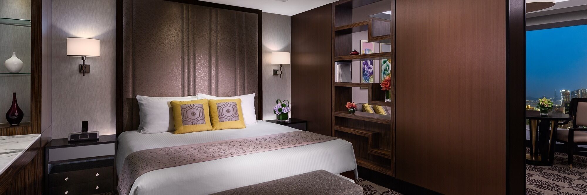 Ein Hotelzimmer mit einem großen Bett, Nachttischlampen, einem Nachttisch, einem Bücherregal und Blick auf die Stadtlandschaft.
