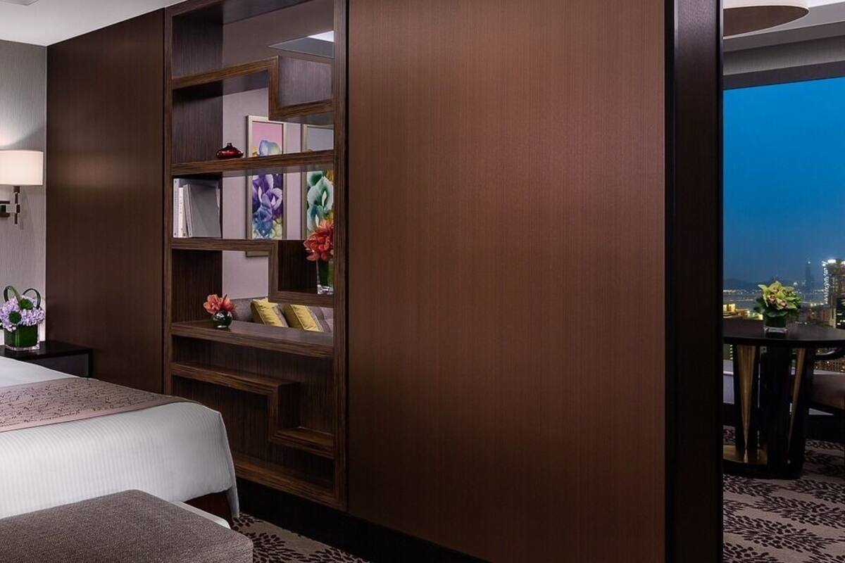 A hotel room with a large bed, bedside lamps, a nightstand, a bookshelf, and a view of the cityscape.