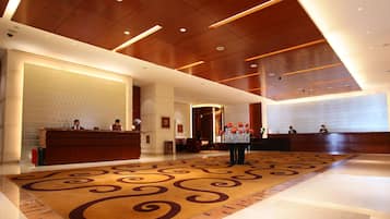 A hotel lobby with a reception desk, two receptionists, a patterned carpet, and a modern ceiling design.