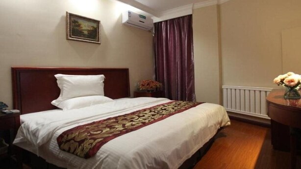 A hotel room with a large bed, a nightstand, a desk, a chair, a window with curtains, an air conditioning unit, and a framed picture on the wall.