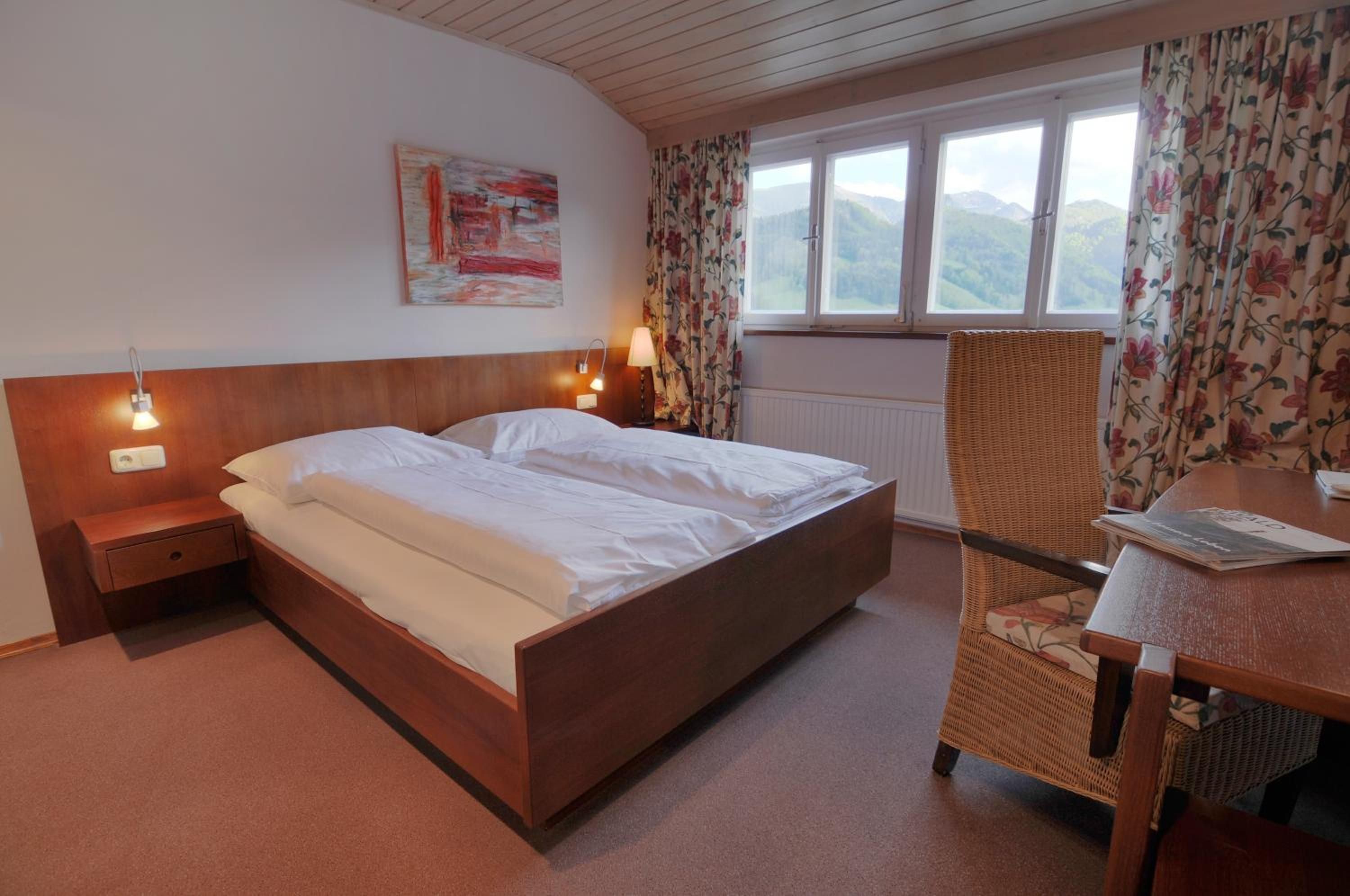 A hotel room with a wooden bed, a nightstand, a lamp, a chair, a table, and a window with a view of mountains.