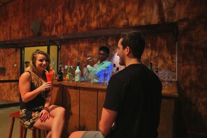A bar area with a wooden interior, a counter with various bottles, and a person seated at the bar. - The Rainforest Ecolodge Sinharaja (Deniyaya)