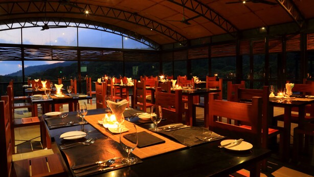 An outdoor dining area with tables set for two, under a curved roof structure with a view of the mountains. - The Rainforest Ecolodge Sinharaja (Deniyaya)