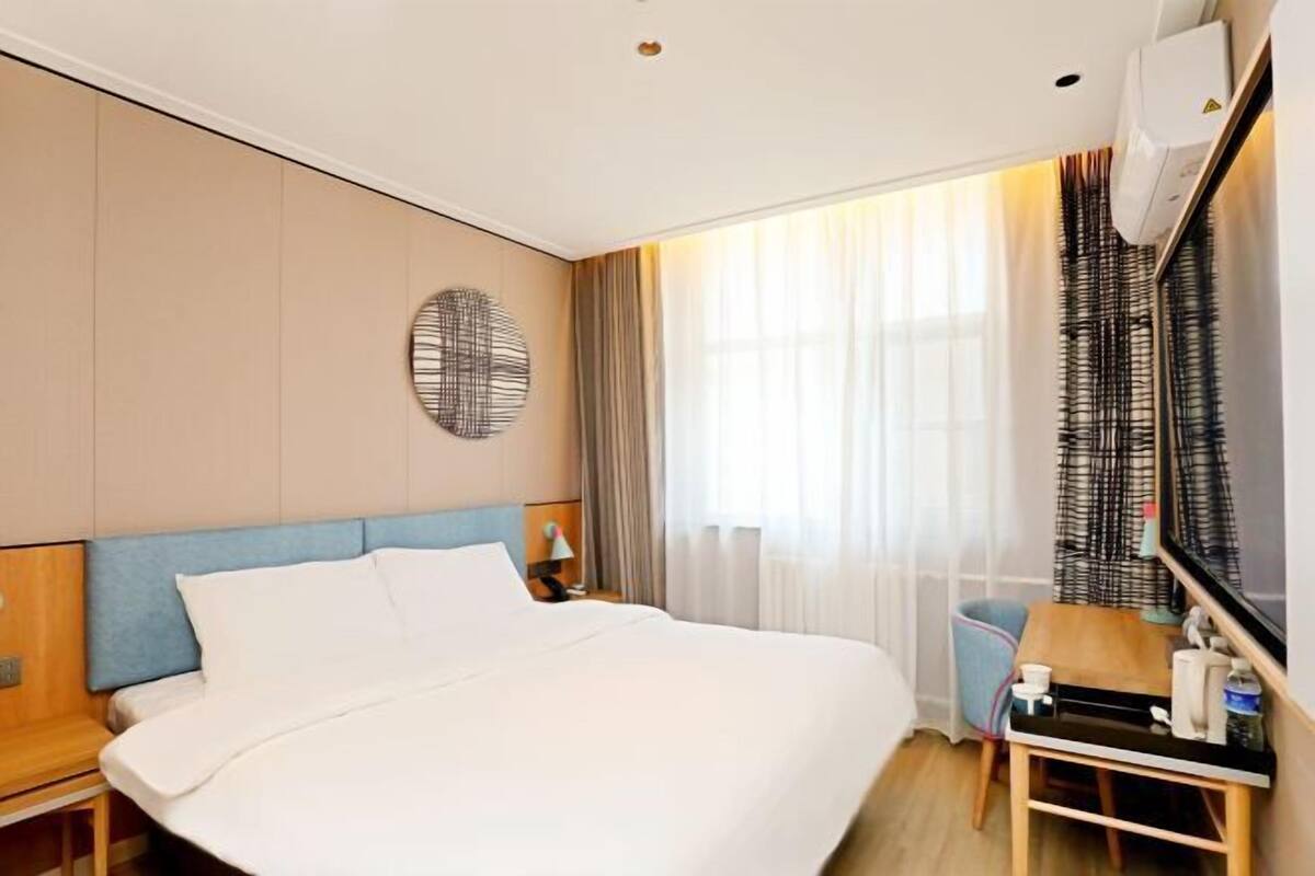 A hotel room with a bed, a desk, a chair, a mirror, a wall-mounted air conditioner, and a window with curtains.