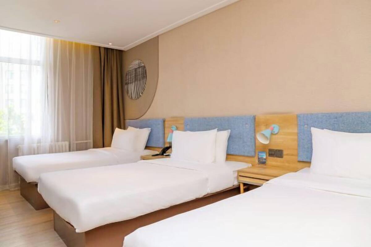 A hotel room with three single beds, a wooden headboard, a bedside table with a lamp, and a large window with curtains.
