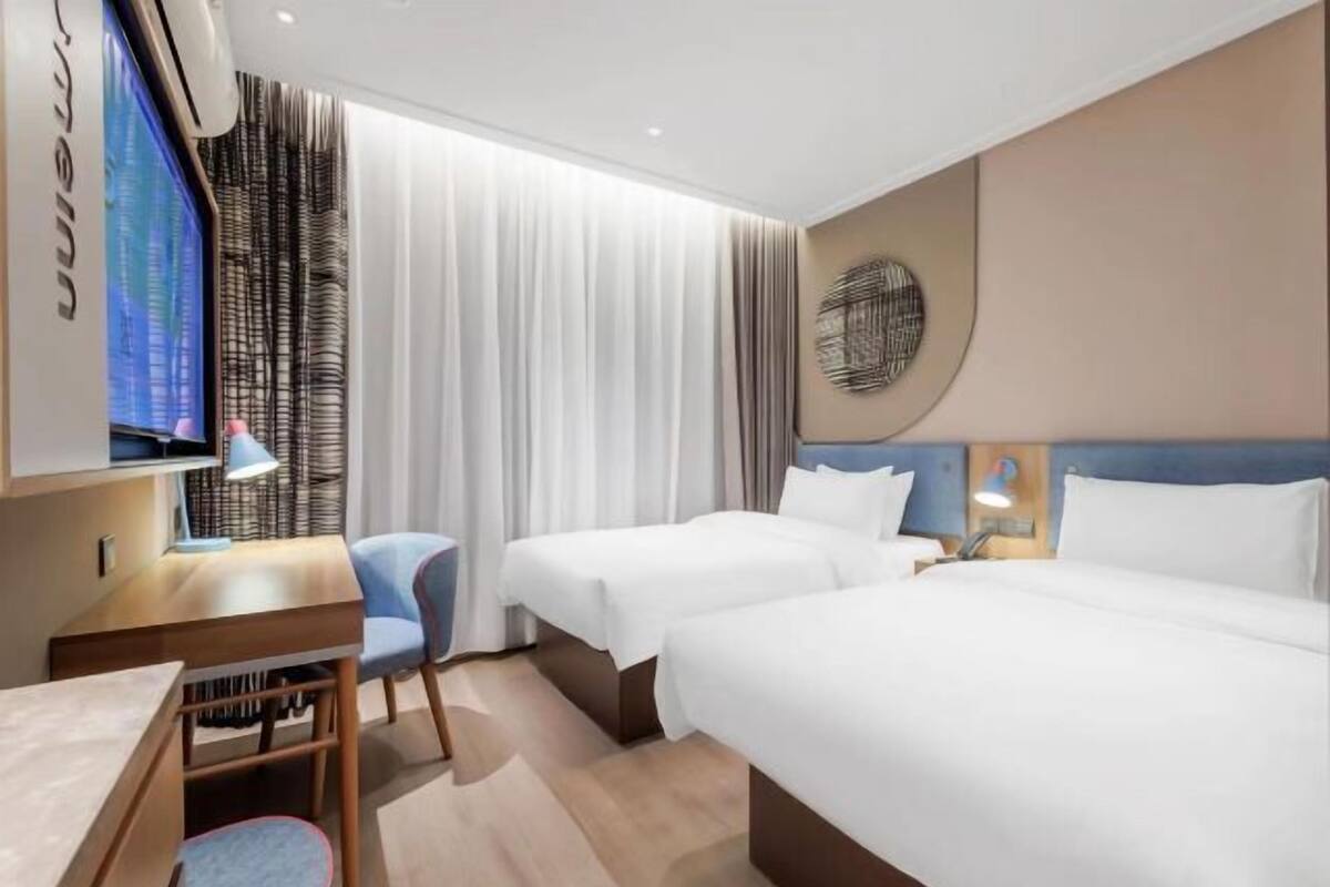 A hotel room with two beds, a desk with a chair, a TV, and a large window with curtains.