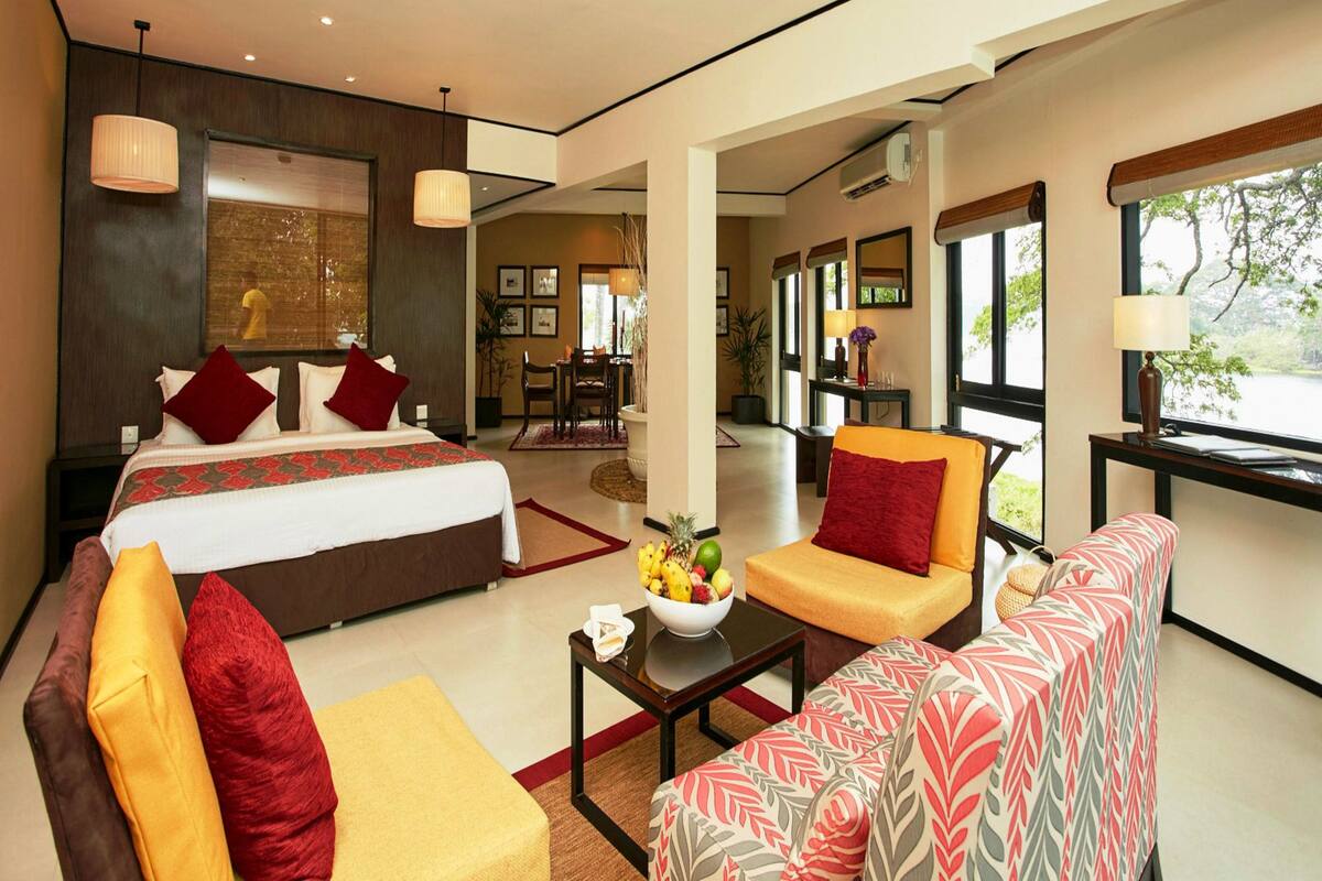 A modern hotel room with a bed, sofa, and armchair, a dining area, and large windows.