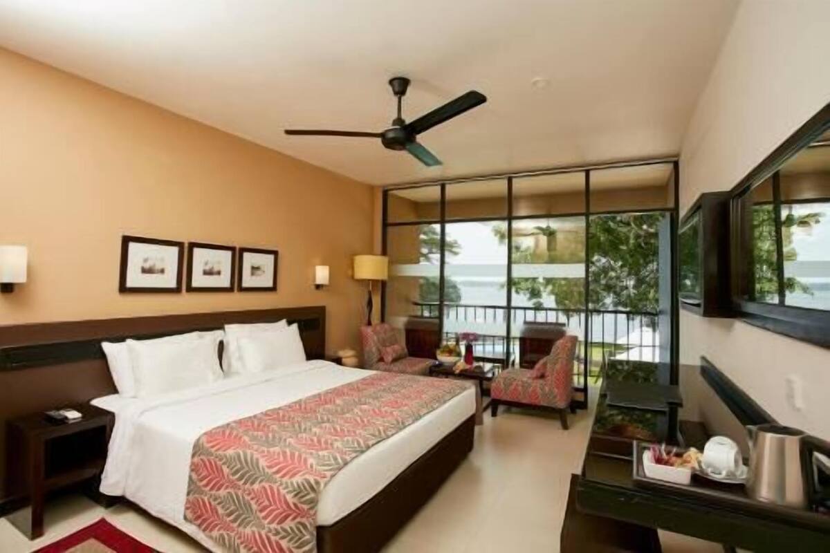 A hotel room with a bed, a desk, a chair, a ceiling fan, and a large window with a view.