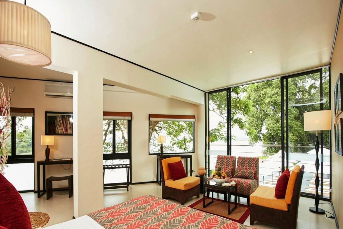 A modern hotel room with a large bed, two armchairs, a small table, and a balcony with a view.