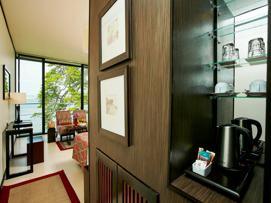 Une chambre d’hôtel moderne dotée d’une grande fenêtre, d’un coin salon avec des fauteuils et une table, d’un espace pour préparer du café et de tableaux encadrés accrochés au mur.