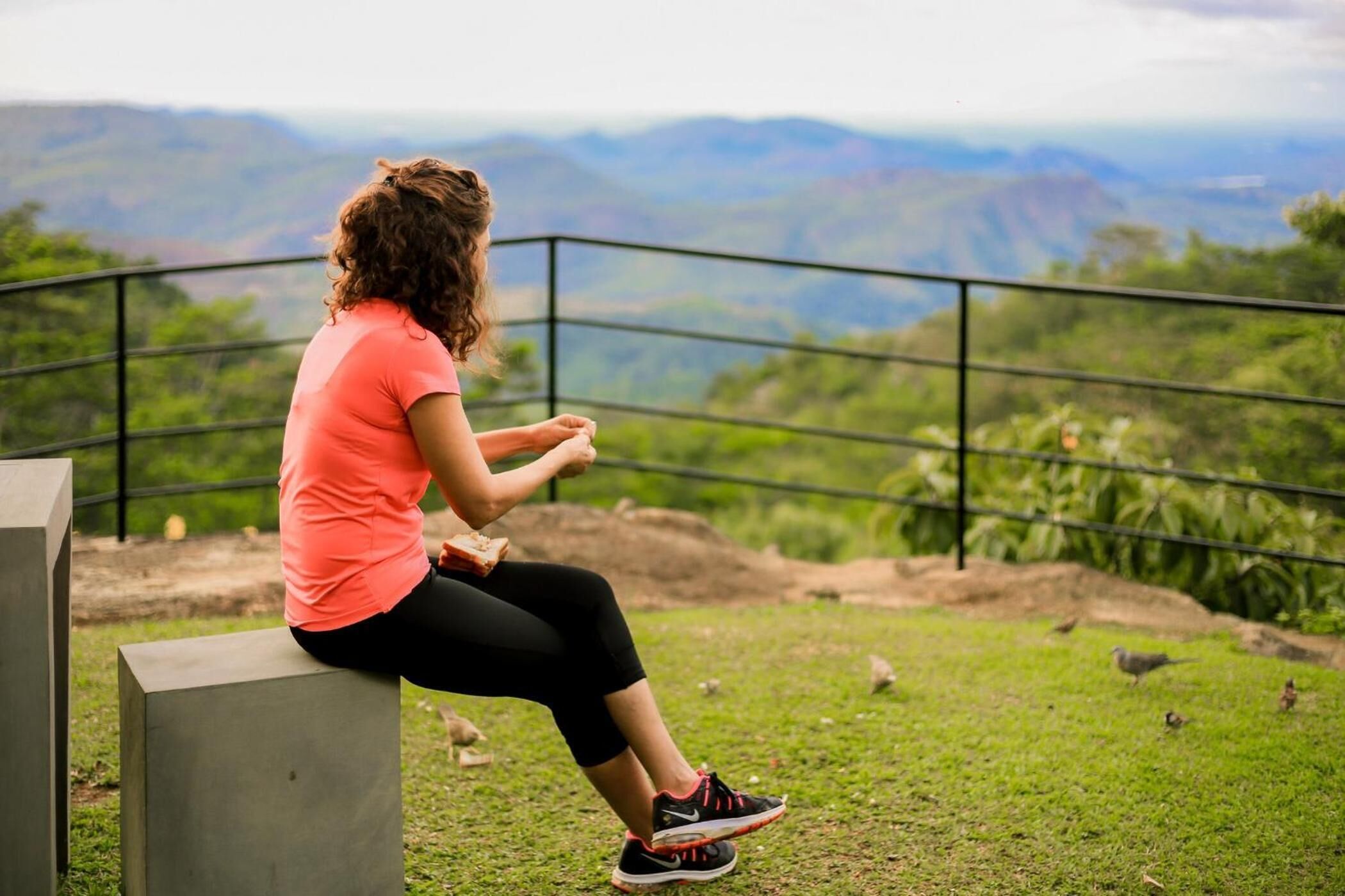 Una persona sentada en un banco de hormigón con vistas a un área verde, una barandilla metálica y colinas distantes.